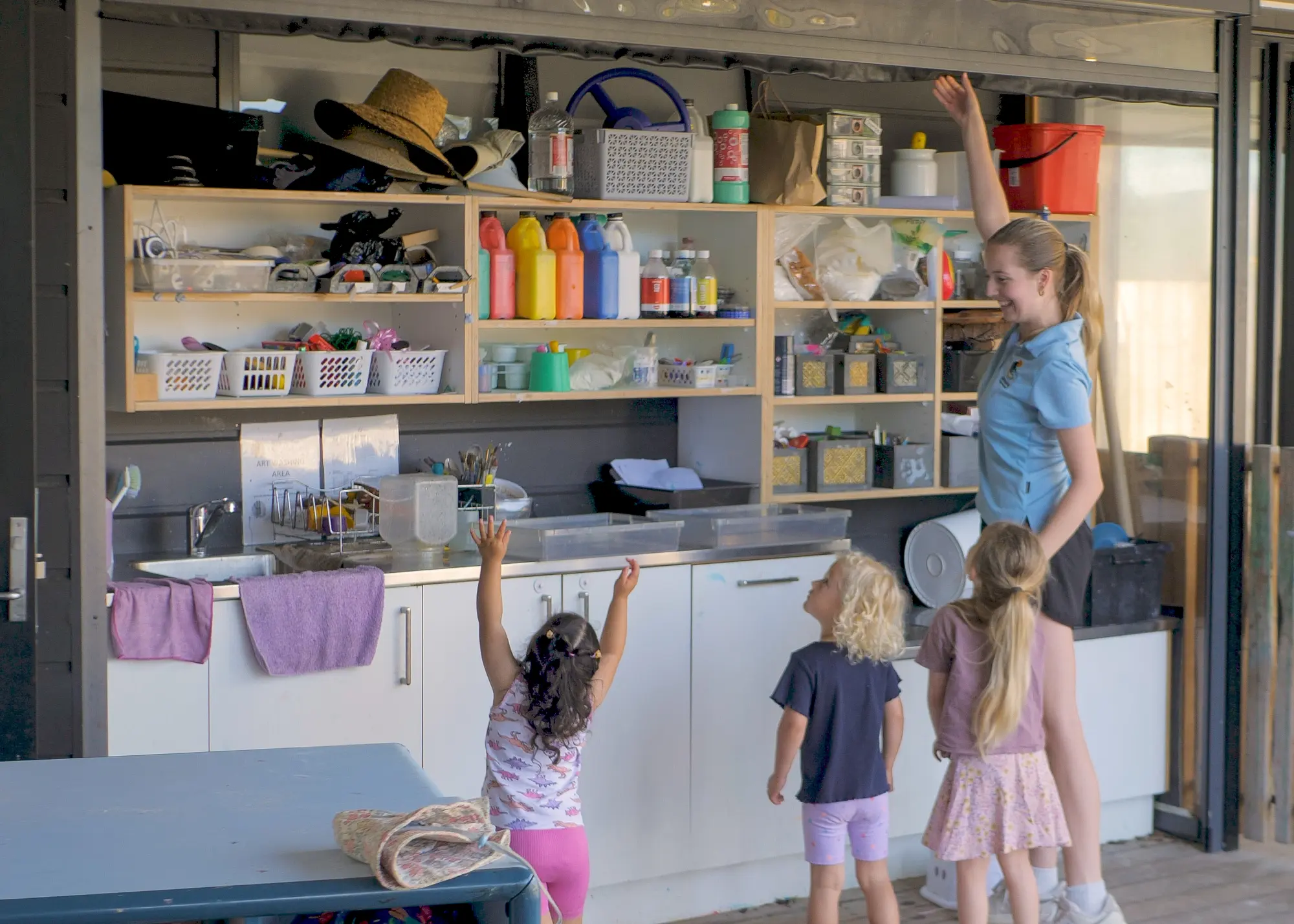 The covered art area with its doors folded open to the courtyard, a teacher and three children reaching up to the paint shelves.