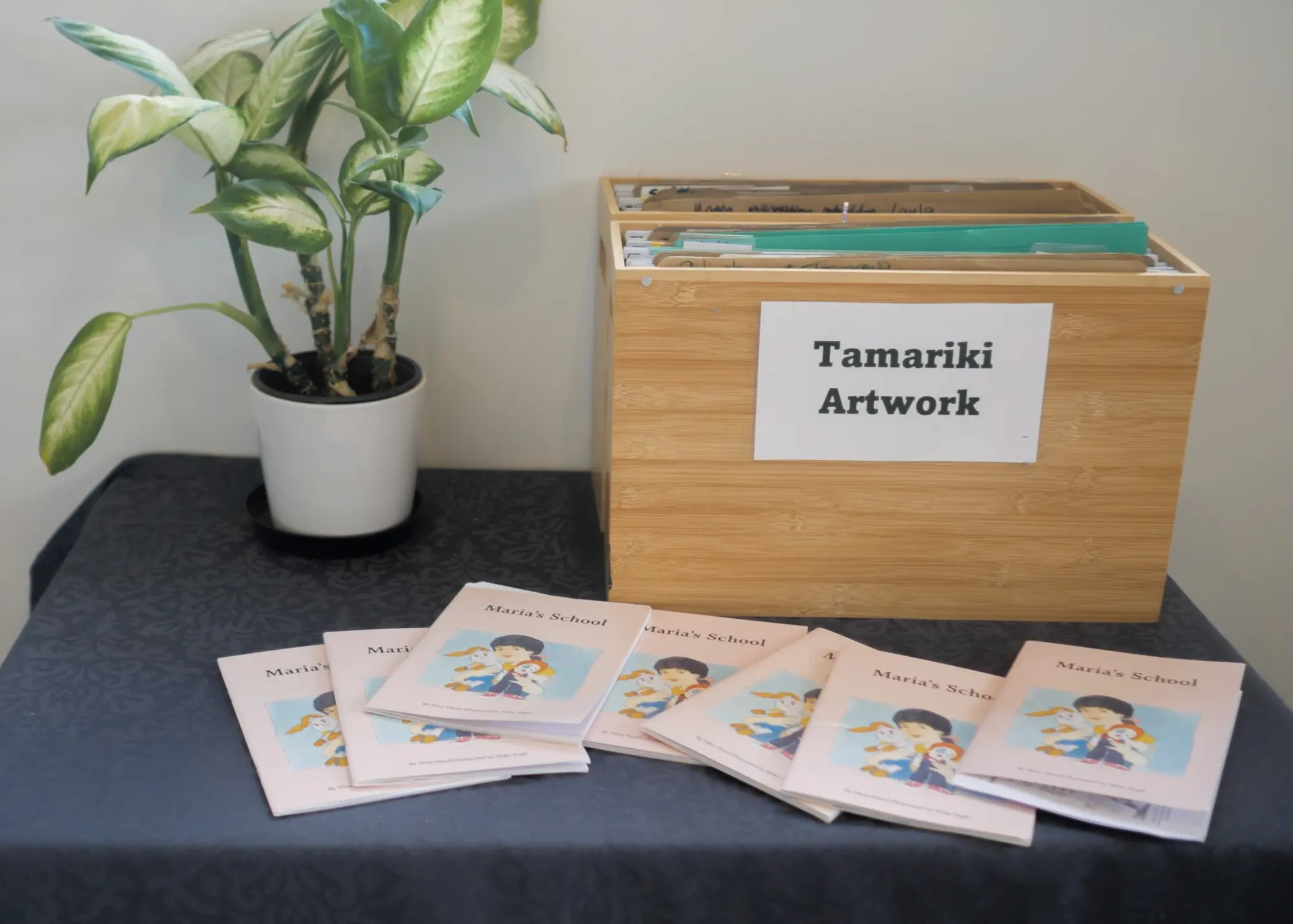 A wooden box labelled Tamariki Artwork on a table beside a pot plant, with children's handmade books laid out in front.