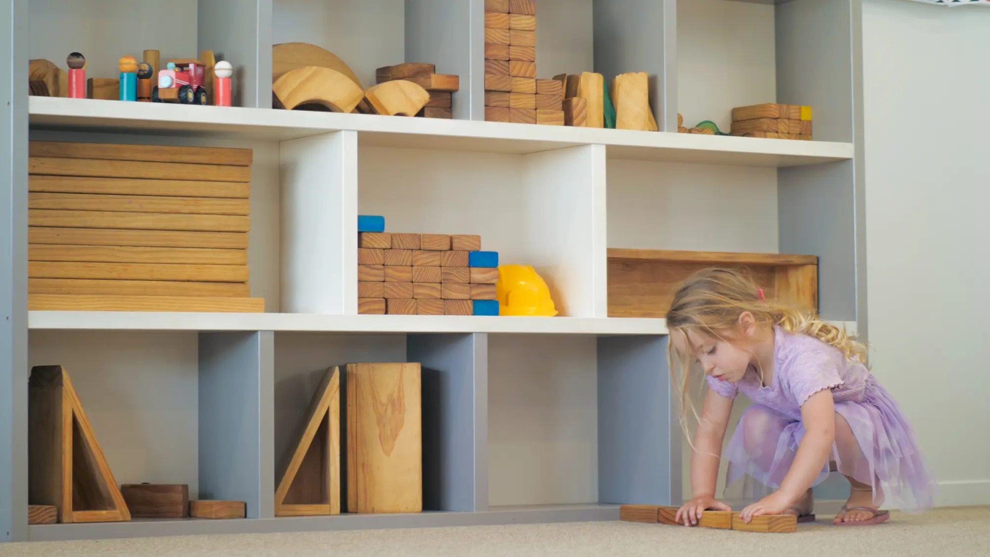 A wall of open shelving filled with wooden blocks, ramps and arches at child height, with a child crouched on the floor building beside it.