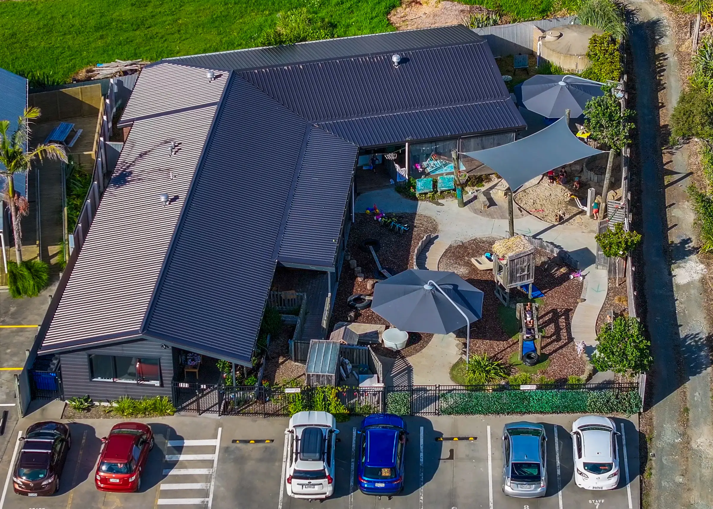 The preschool from above: its dark roof, the shade sails and umbrellas over the sandpit and play area, children playing outside, and the carpark along the front.