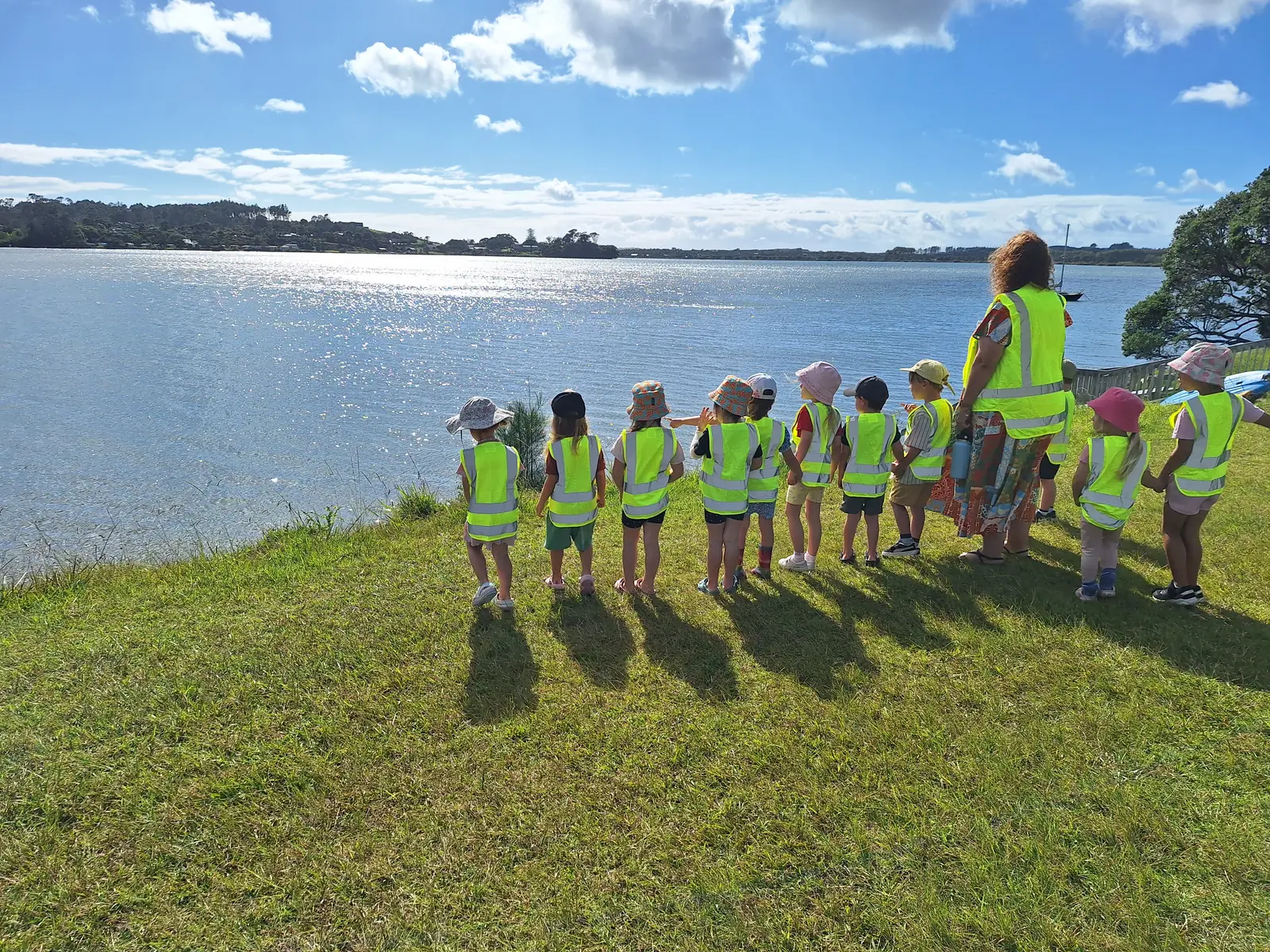 A line of children in hi-vis vests standing with their teacher on the grass, looking out over the Mangawhai estuary.