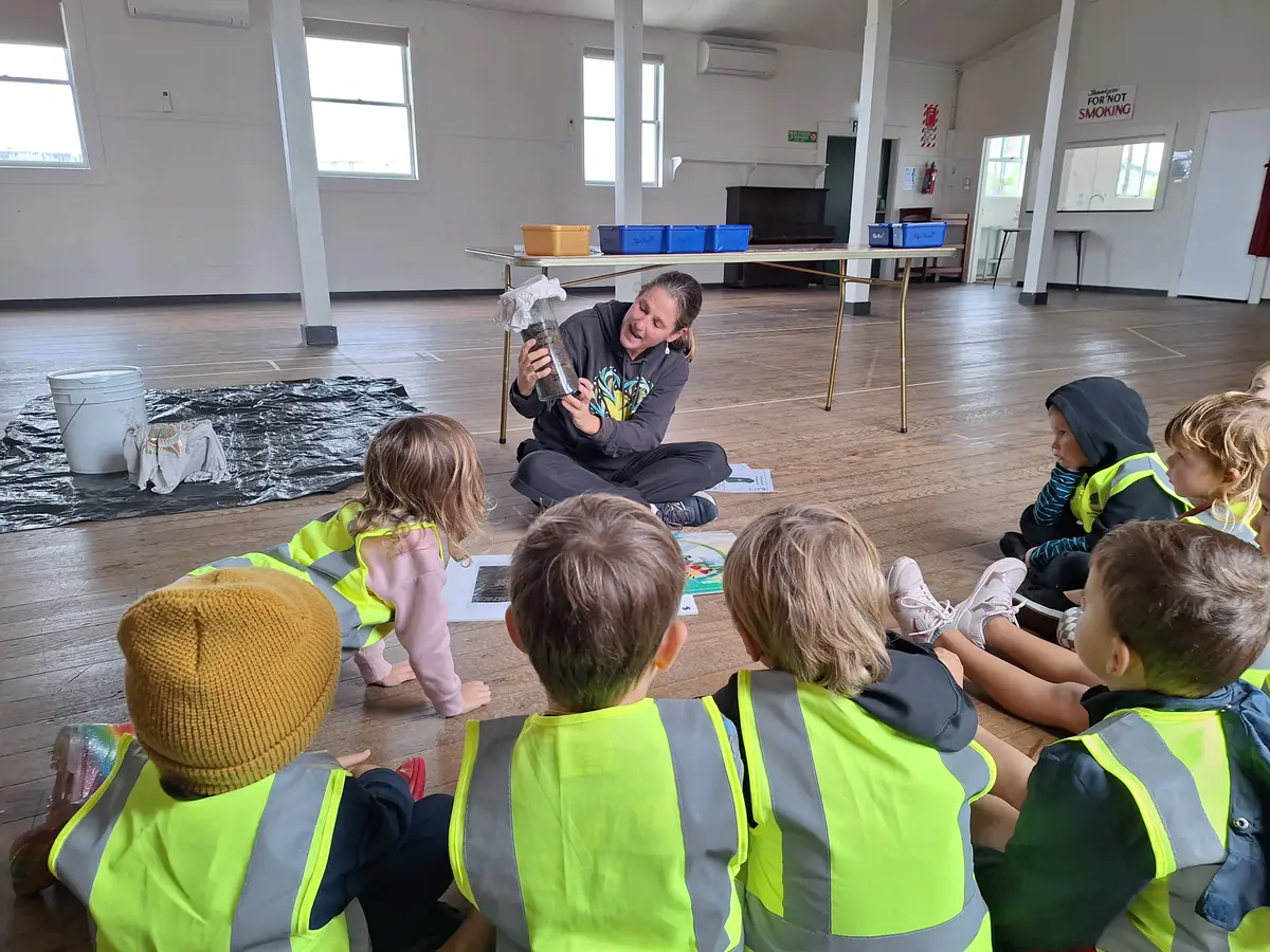A teacher sitting on the floor of the library hall reading to a group of children in hi-vis vests.