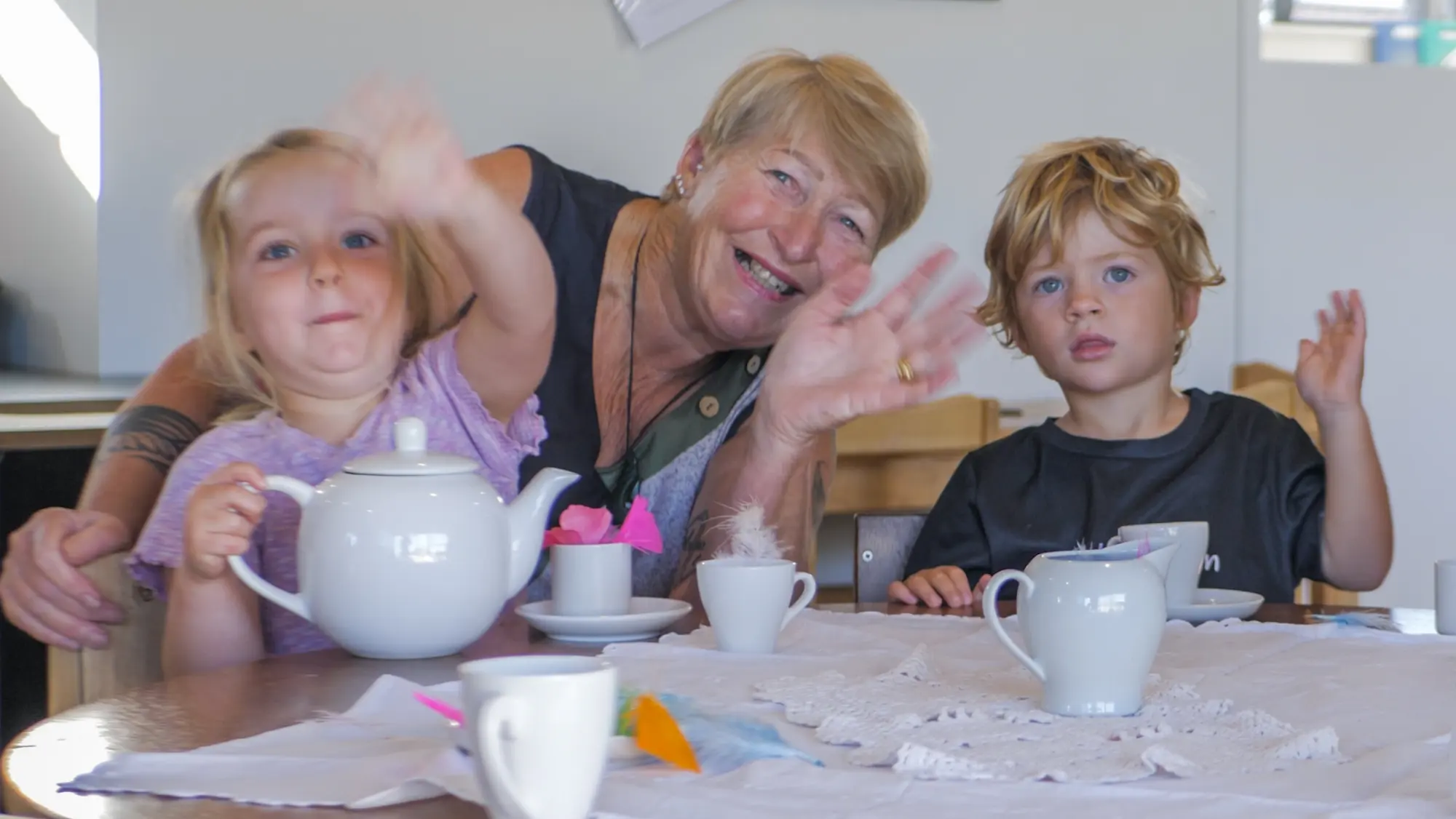A teacher and two children at a table set with a teapot and cups, all waving at the camera.