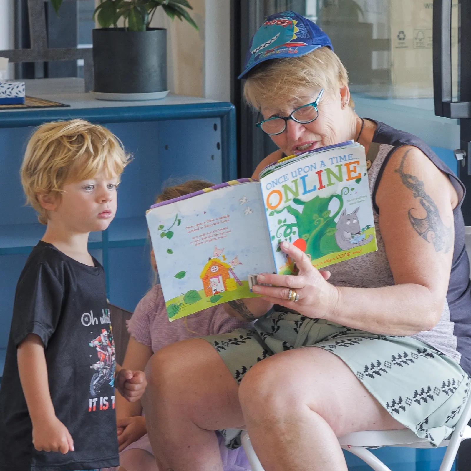 A teacher in a cap and glasses reading a picture book aloud, with a child standing beside her looking on.