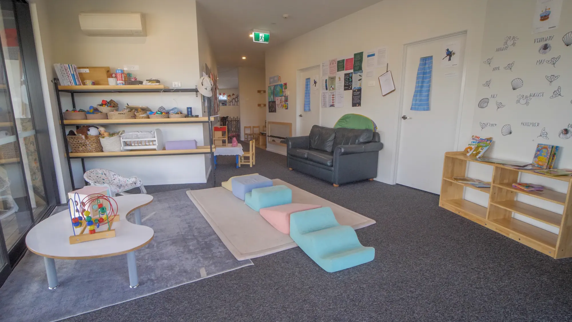 An indoor room with a low table, soft play shapes on a rug, a couch and open shelves of books and baskets.