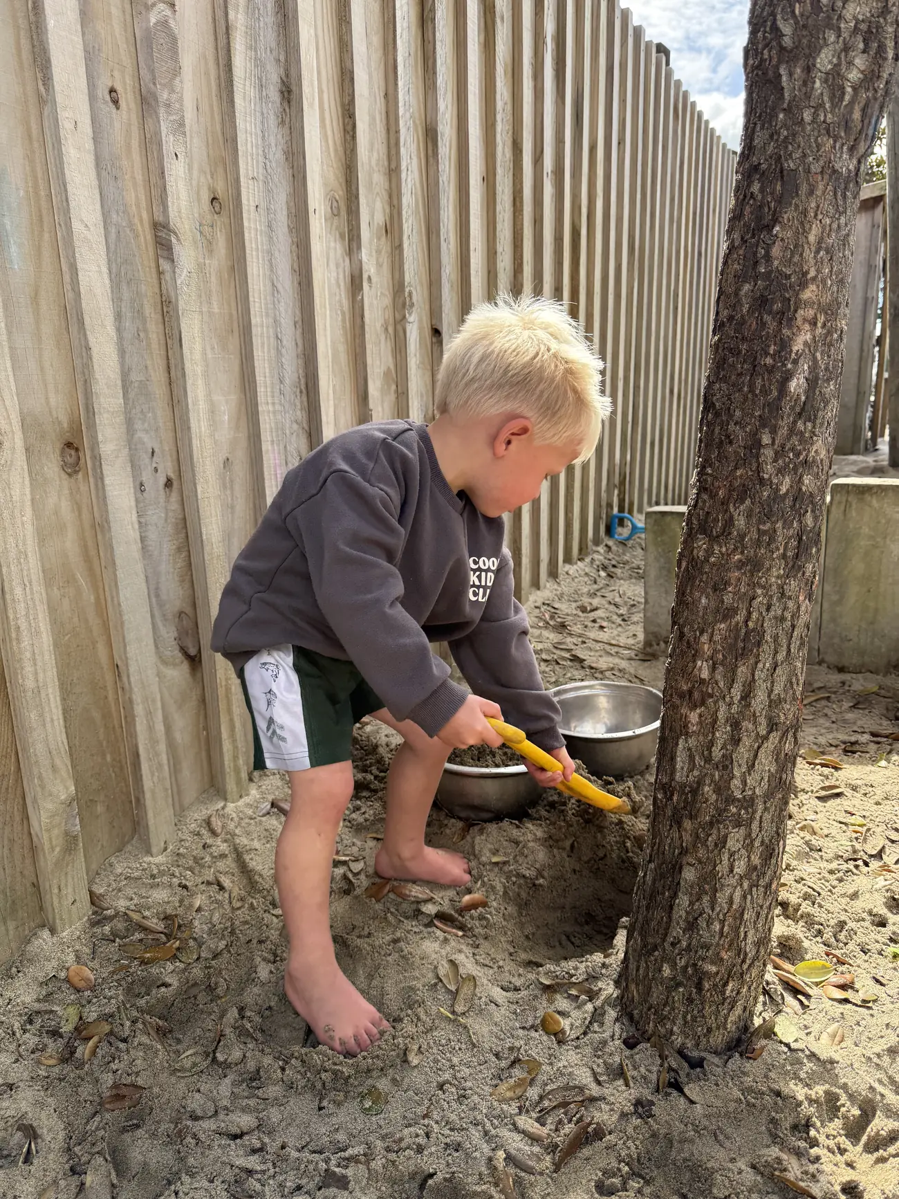 A child digging a hole in the sandpit with a yellow spade beside a tree and the timber fence.