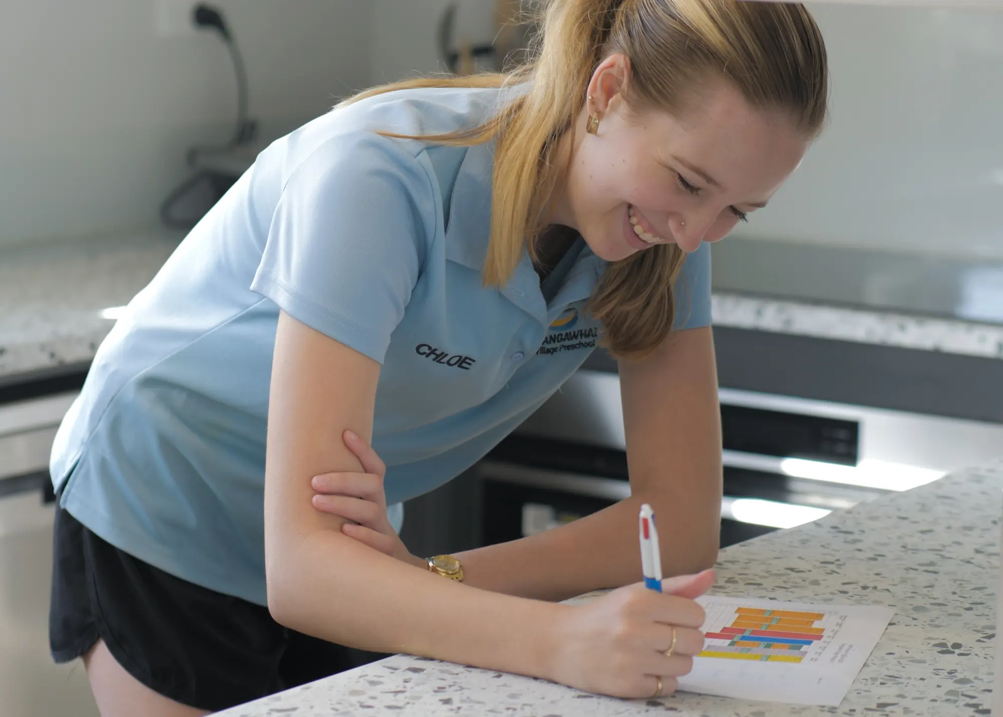 A teacher in a Mangawhai Village Preschool polo leaning on the kitchen bench, smiling as she fills in a colour-coded chart.