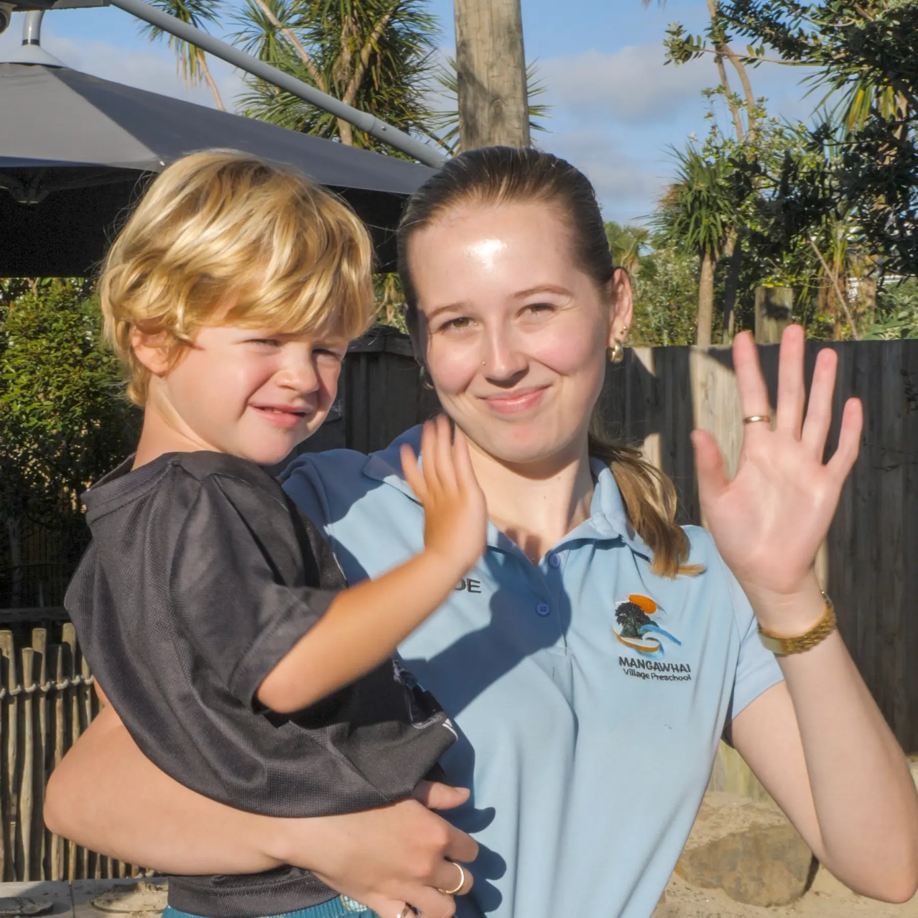 A teacher in a Mangawhai Village Preschool polo shirt carrying a child, both waving at the camera.