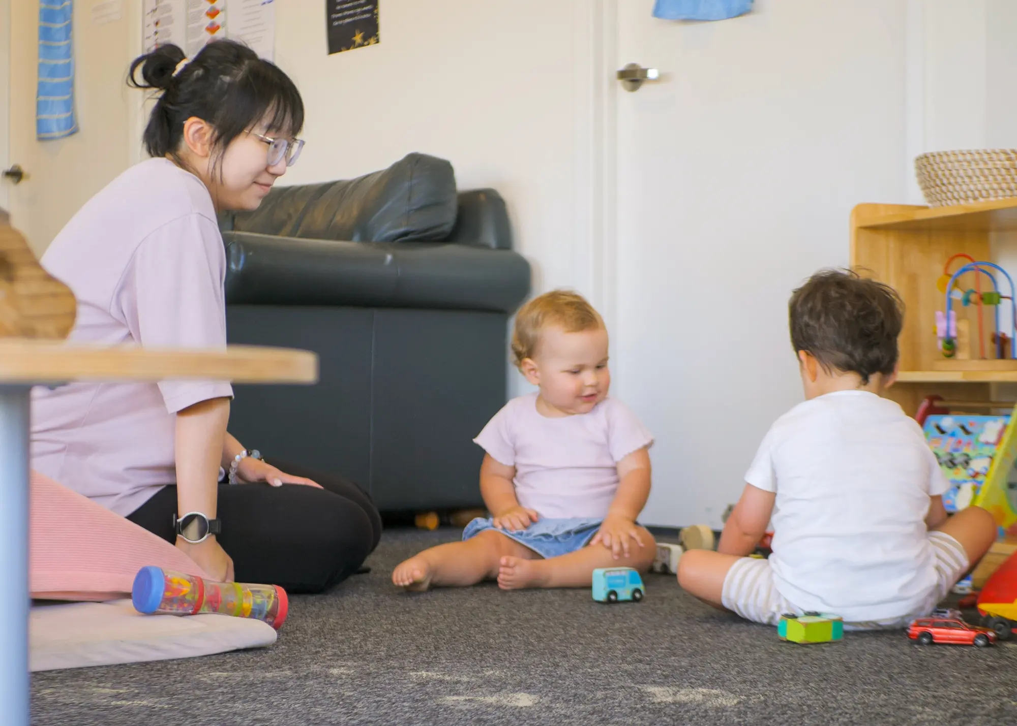A kaiako sitting on the floor of the under twos room with two toddlers playing with wooden cars beside a low shelf of toys.