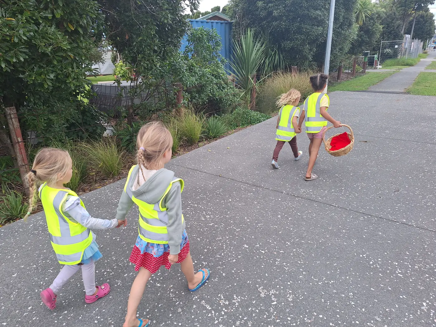 Four children in hi-vis vests walking along the footpath in Mangawhai village, one carrying a basket.
